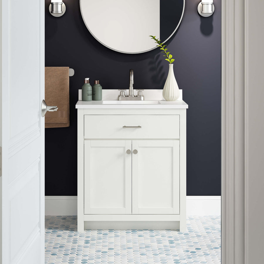 A modern bathroom with a white vanity, round mirror, dark walls, and patterned tile floor. A vase with greenery, toiletries, and a brown towel add decorative touches. The sink faucet is centered on the vanity.