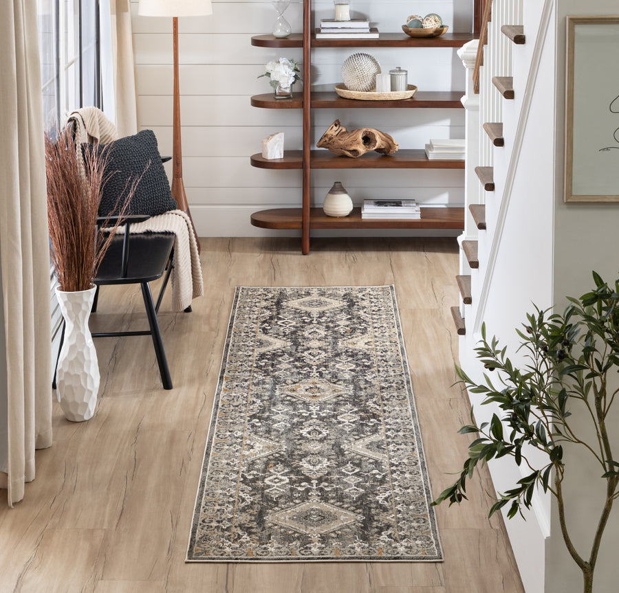 A stylish hallway features a patterned runner rug on light wood floors, a black chair with a cushion, shelves with decorative items, a potted plant, and stairs with a white railing. Natural light streams in from the left.