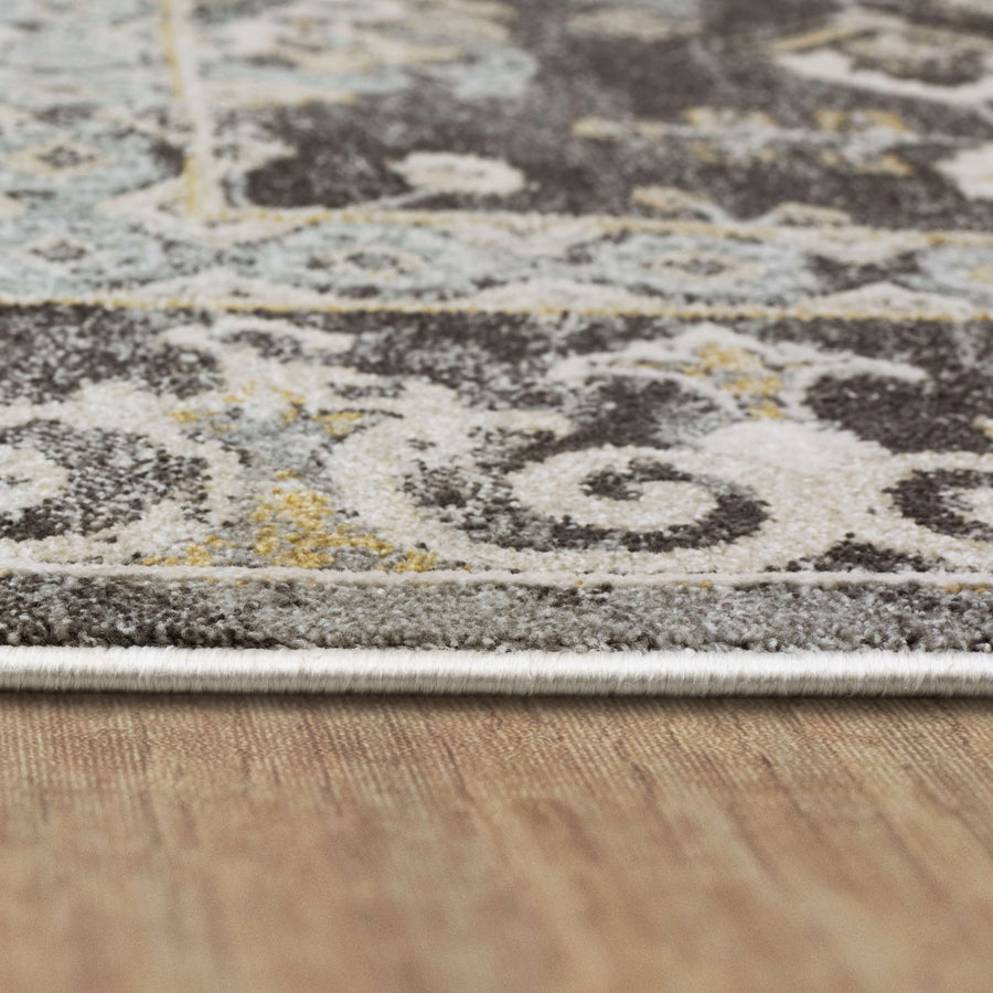 Close-up of the edge of a patterned area rug with gray, white, and yellow tones, lying on a wooden floor. The rug has a slightly distressed, intricate design.