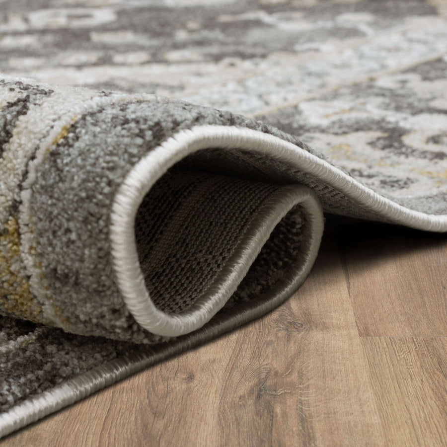 A close-up of a gray and beige patterned rug with its edge folded over on a wooden floor, showing the texture and thickness of the carpet.
