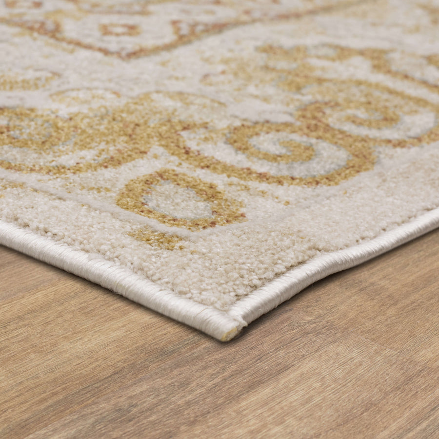 Close-up of a beige patterned rug with gold and brown swirls, placed on a wooden floor. The rugs edge is neatly finished with white binding.