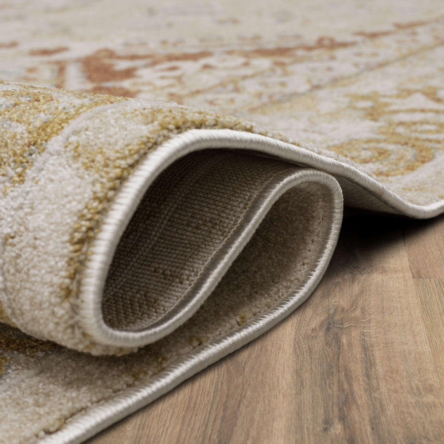 A close-up view of a beige and gold patterned rug partially folded over on a wooden floor, showing the rug’s textured surface and stitching along the edge.