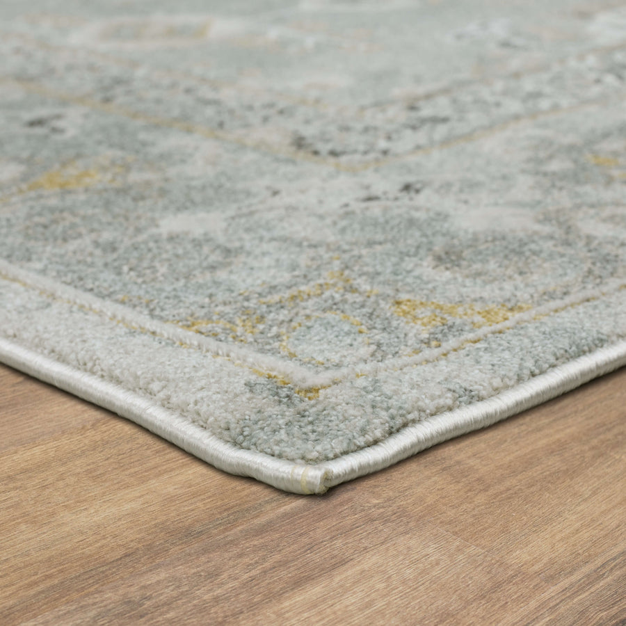 Close-up view of the corner of a patterned area rug with muted blue, grey, and yellow tones on a wooden floor. The rug has a soft texture and a neatly finished edge.