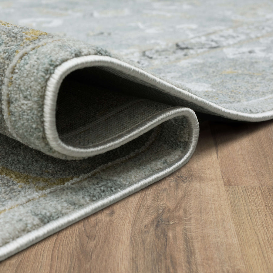 A close-up of a light-colored area rug with gray and beige tones, partially folded over itself, resting on a wood floor.