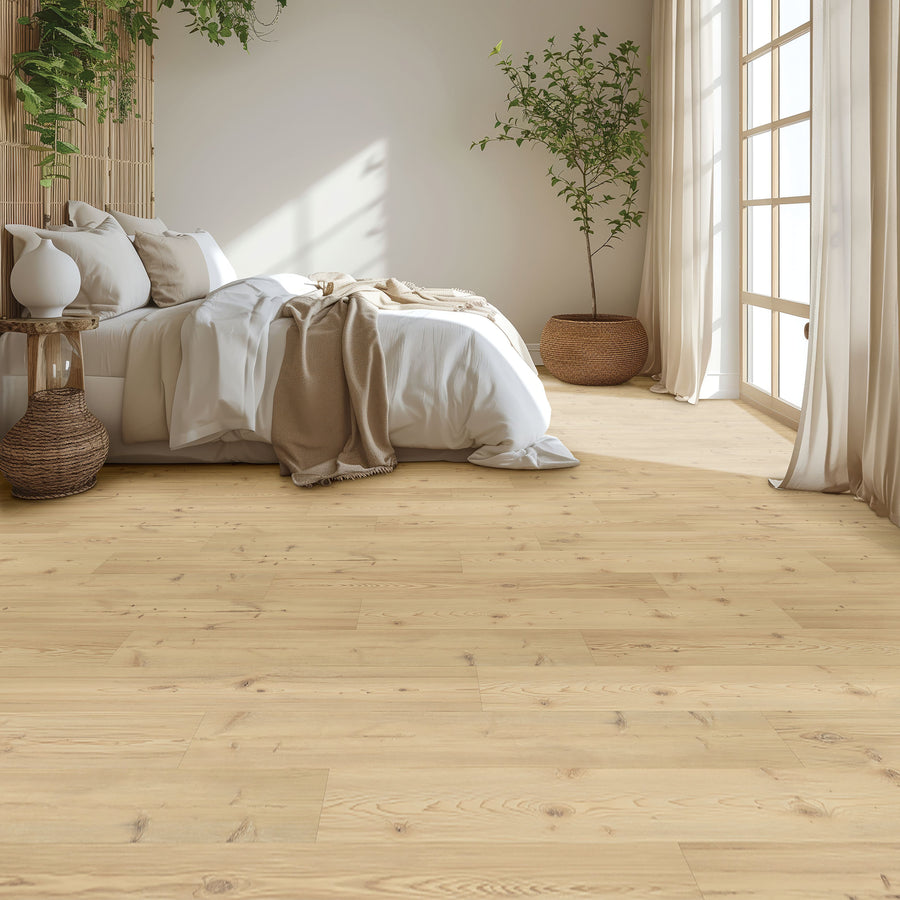 A cozy bedroom featuring Glacial Isle 8 11/32" Waterproof Laminate flooring, a white bed with beige blankets, a potted tree, wicker basket, and large windows allowing sunlight through sheer curtains.