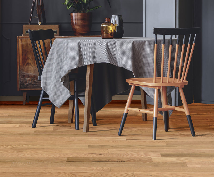 A dining table with a gray tablecloth is set with glass vases and jars. Two wooden chairs with black spindle backs are positioned on opposite sides, on a light wood floor in a cozy, modern room.
