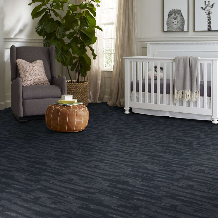 A cozy nursery with a gray armchair, a brown leather pouf, a crib with a gray blanket, a large potted plant, and animal artwork on the wall. The room has dark blue patterned carpet flooring.