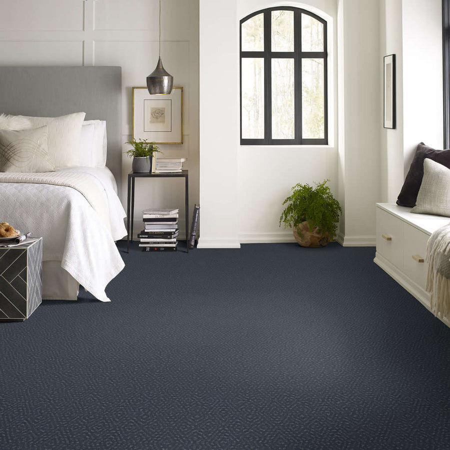 Modern bedroom with a gray upholstered bed, white bedding, black nightstand, hanging lamp, large window, green plant, and dark gray carpet flooring. Light walls and built-in seating create a cozy, airy atmosphere.