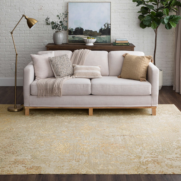 A cozy living room with a light beige sofa adorned with cushions and a throw blanket, set on a patterned beige rug. Behind the sofa is a console table with decor, artwork, and a tall green plant in the corner.