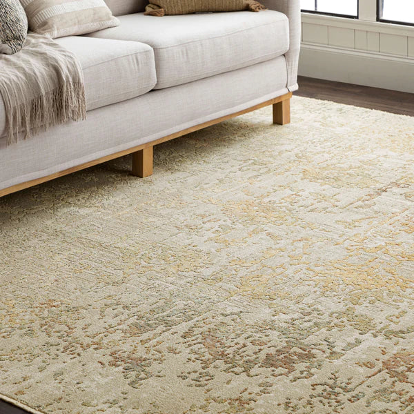 A light-colored, textured area rug with subtle patterns is placed in front of a beige sofa with wooden legs and neutral-toned pillows, next to a window letting in natural light.