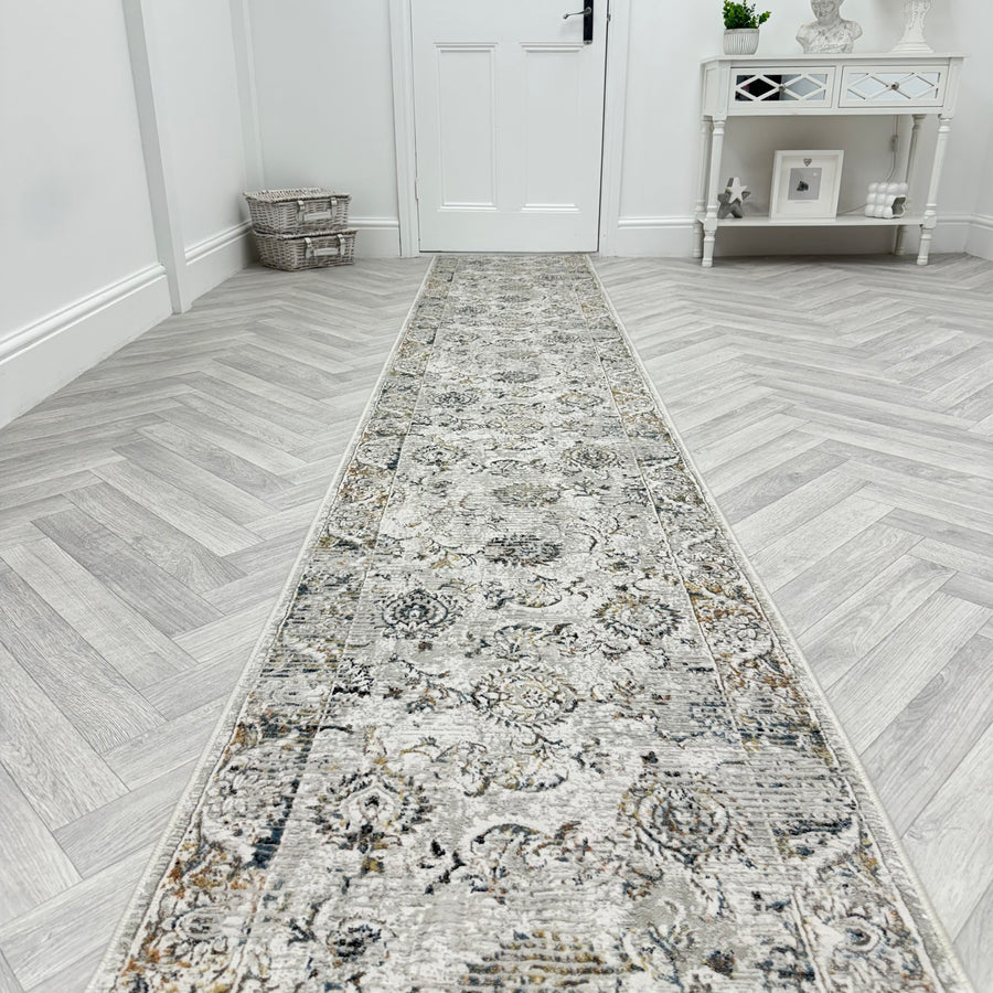 A hallway with light herringbone floors showcases a Reha 1061-199 Runner Rug leading to a white door. Against the right wall, a decorative white console table stands below a mirror with a basket beside it.