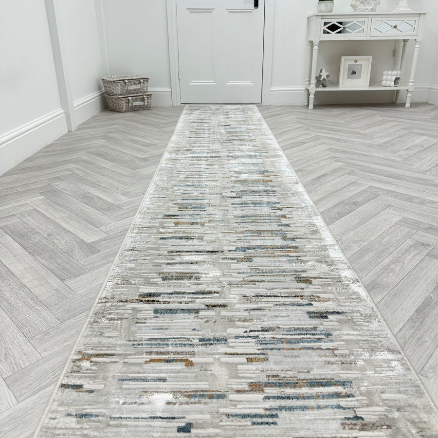 The Reha 1064-190 Runner Rug stretches down a hallway with light gray herringbone floors, ending at a white door. A small white table with decor and a woven basket on the left complete the modern, minimalist look.