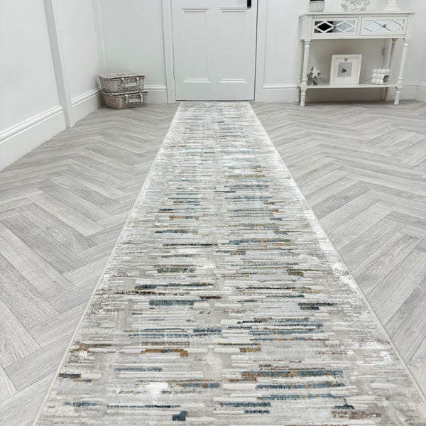 The Reha 1064-190 Runner Rug stretches down a hallway with light gray herringbone floors, ending at a white door. A small white table with decor and a woven basket on the left complete the modern, minimalist look.