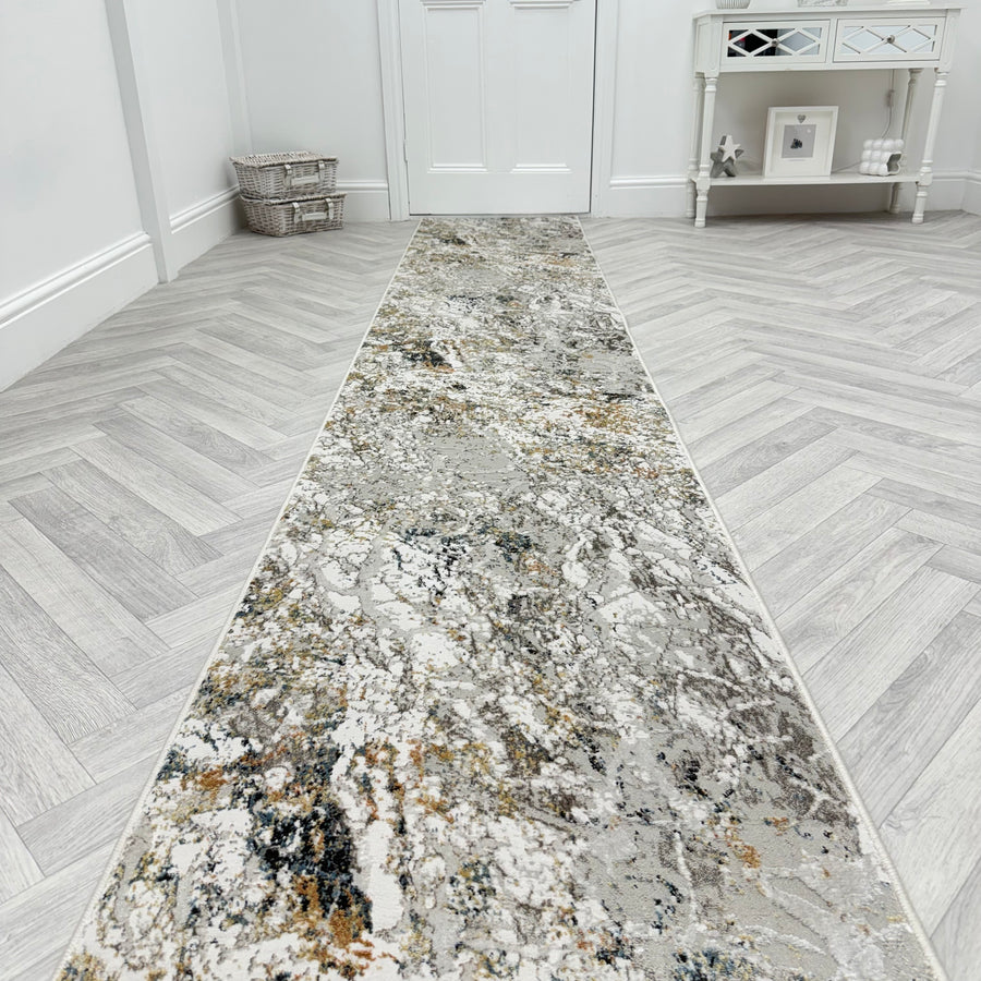 The Reha 1065-199 Runner Rug, featuring abstract gray, beige, and gold tones, lies on a light herringbone floor in a bright hallway with white walls, a basket, and a mirrored console table holding decor.