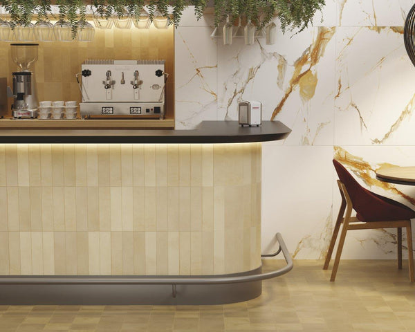 A modern coffee bar with a light wood counter, an espresso machine, stacked cups, hanging glasses, and a marble-patterned wall. A round table with two chairs is partially visible to the right.