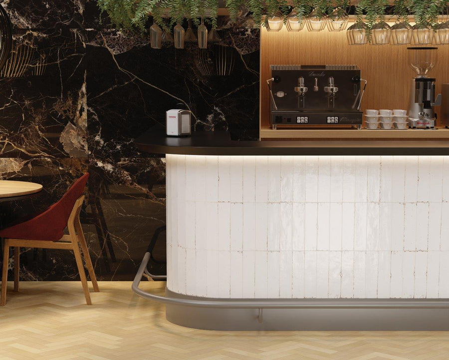 A modern coffee bar with a white tiled counter, espresso machine, stacked cups, and a black marble wall. Part of a round table with a red velvet chair is visible on the left. Green foliage hangs above.