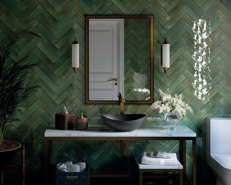 Modern bathroom with green herringbone tile wall, a rectangular mirror, two wall sconces, a black vessel sink on a white countertop, decorative baskets, flowers, and neatly folded towels.