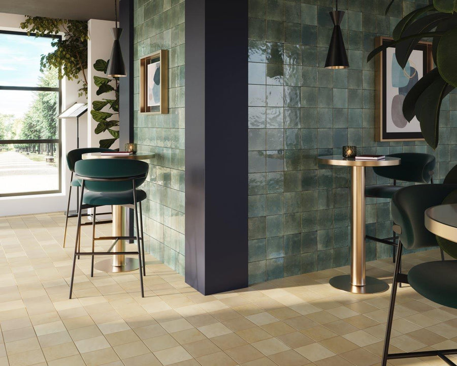 Modern cafe interior with teal tiled walls, gold-accented round tables, dark green chairs, framed art, hanging pendant lights, plants, and large windows letting in natural light.