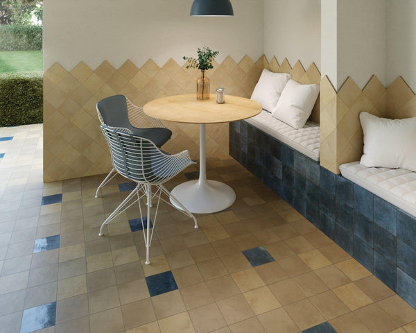 A cozy dining nook with a round wooden table, two modern chairs, a built-in cushioned bench with pillows, and geometric beige and blue tile flooring and walls. A plant sits on the table under a pendant light.