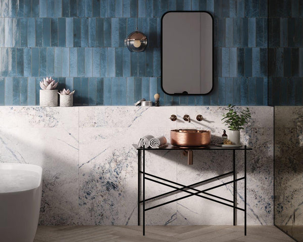 A modern bathroom with blue vertical wall tiles, a marble backsplash, a rectangular mirror, a round copper sink on a minimalist metal stand, potted plants, rolled towels, and a bathtub partially visible on the left.