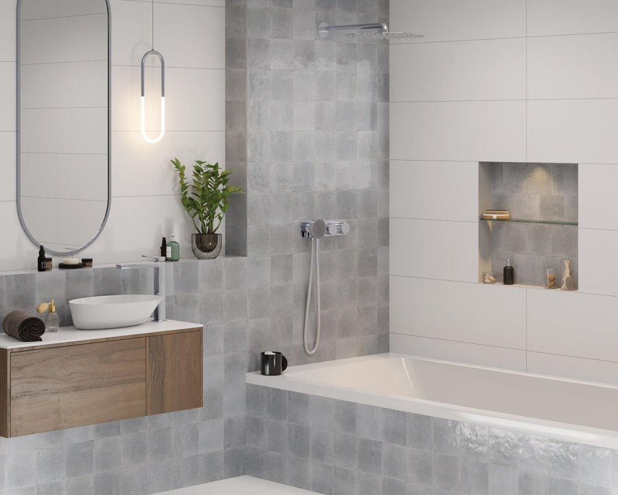 Modern bathroom with gray and white tiles, a built-in bathtub, wall-mounted sink with wood cabinet, oval mirror, small potted plant, and recessed shelf holding toiletries. Soft lighting creates a clean, minimalist look.