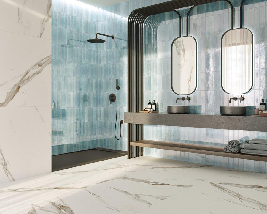 Modern bathroom with marble floors and walls, a blue-tiled shower area with black fixtures, two large mirrors, double sinks on a concrete counter, and towels neatly stacked on a wooden shelf below.