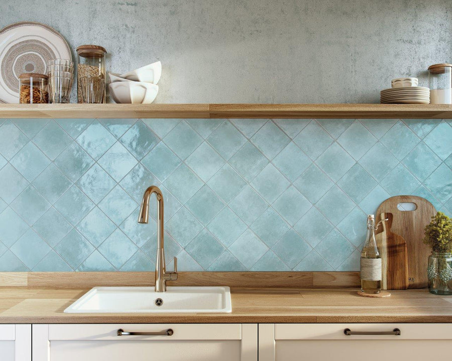 A modern kitchen countertop with a white sink and a gold faucet, light wood surfaces, blue diamond-patterned tile backsplash, and open shelves holding dishes, jars, and kitchen decor.