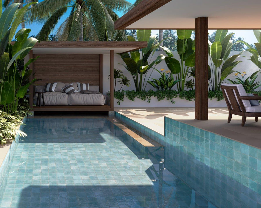 A serene outdoor pool with clear blue water, a modern covered lounge area with a cushioned daybed, tropical plants, and palm trees in the background under bright daylight.