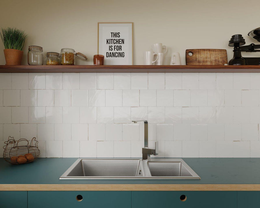 A modern kitchen with a turquoise countertop, a double sink, white tiled backsplash, and a wooden shelf holding jars, mugs, plants, and a framed sign that reads, THIS KITCHEN IS FOR DANCING.