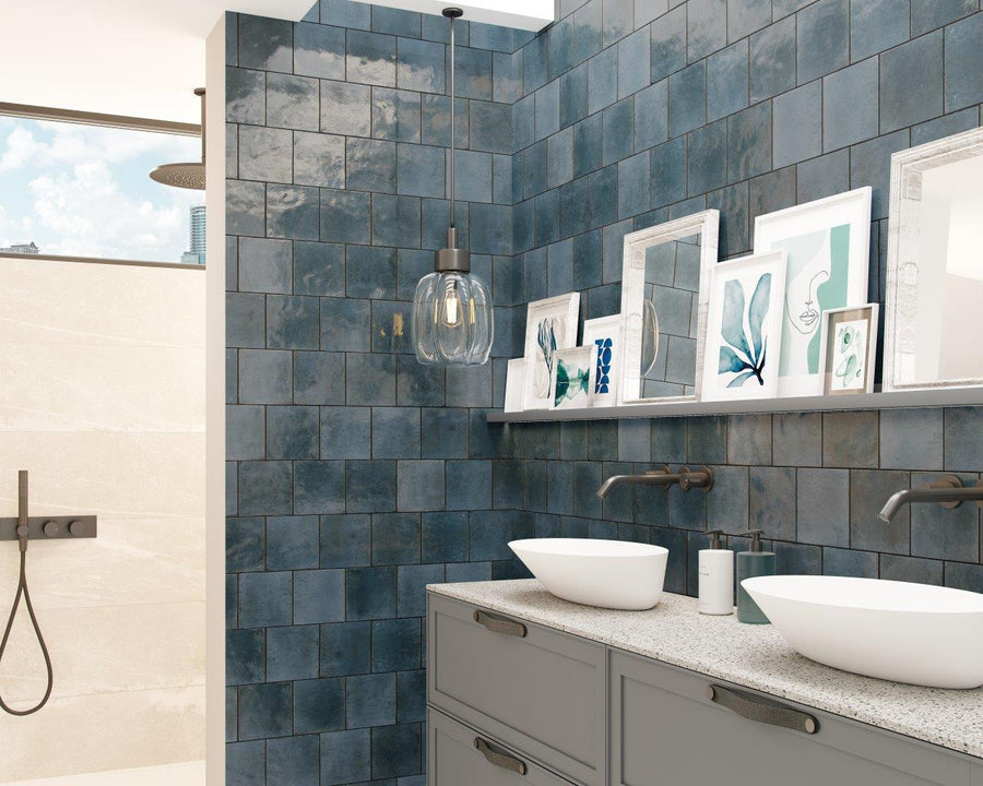 Modern bathroom with blue square wall tiles, a double sink vanity with vessel sinks, wall-mounted faucets, framed art on a shelf, a pendant light, and a walk-in shower with a rainfall showerhead in the background.