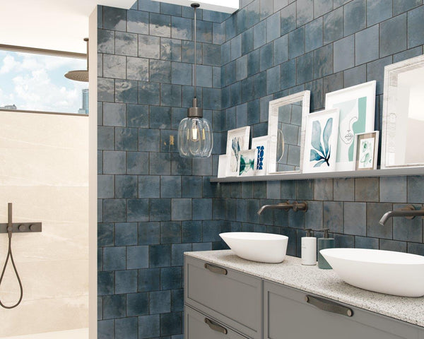 Modern bathroom with blue square wall tiles, a double sink vanity with vessel sinks, wall-mounted faucets, framed art on a shelf, a pendant light, and a walk-in shower with a rainfall showerhead in the background.