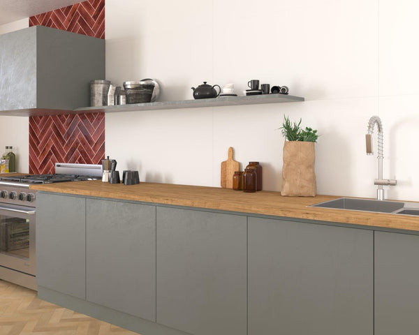 A modern kitchen with gray cabinets, a wooden countertop, red herringbone tile backsplash, open shelf with dishes, a sink, and a brown paper bag with fresh herbs.
