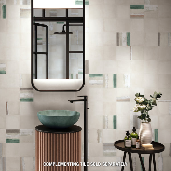 A modern bathroom with a round teal vessel sink, black faucet, wood-textured vanity, round mirror, and a small table holding flowers and toiletries. The wall features a geometric tile pattern with green and beige accents.