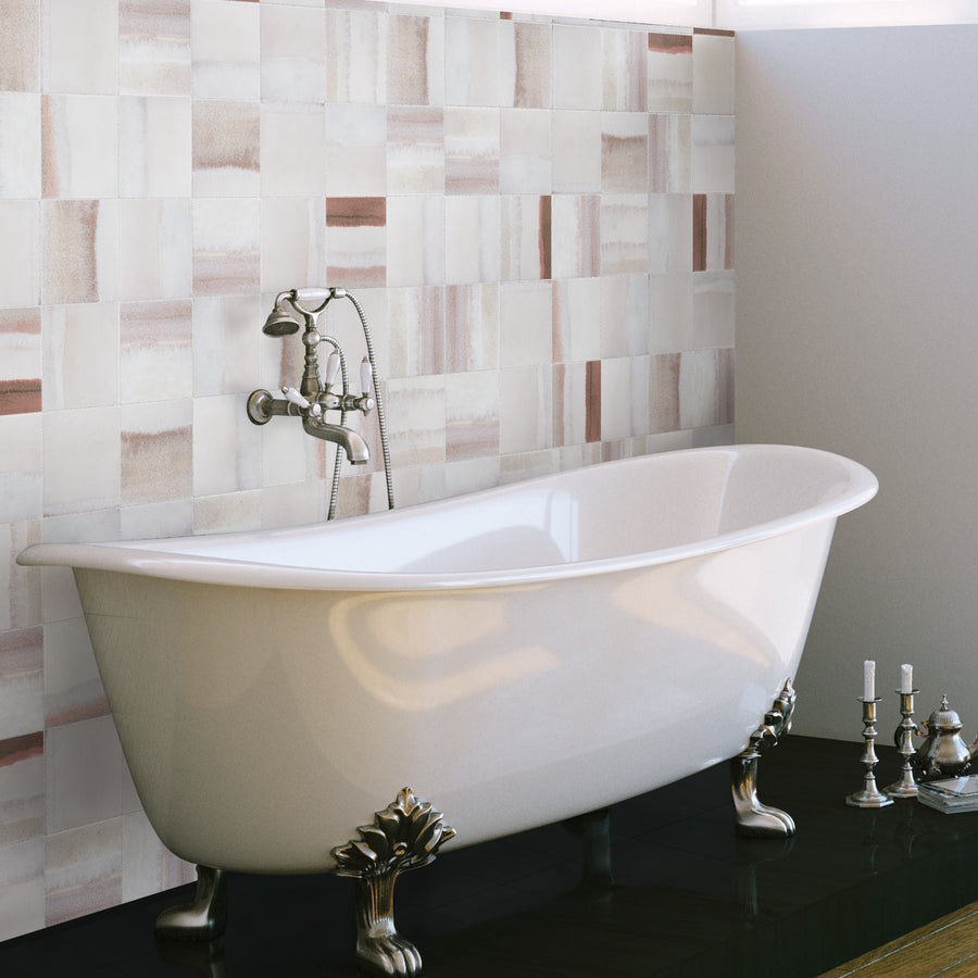 A white clawfoot bathtub with ornate metal feet sits on a dark floor, against a wall with rectangular, beige and pinkish tiles. Nearby, a set of silver candlesticks is placed on the floor.