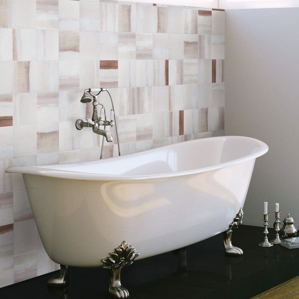 A white clawfoot bathtub with ornate metal feet sits on a dark floor, against a wall with rectangular, beige and pinkish tiles. Nearby, a set of silver candlesticks is placed on the floor.
