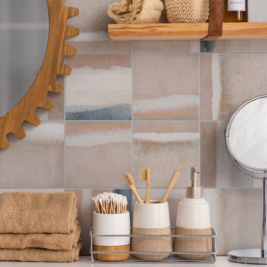 A bathroom counter with beige towels, a wire basket holding cotton swabs, bamboo toothbrushes, and soap dispensers. A round mirror and wooden shelf with rolled towels are visible against patterned tiled walls.