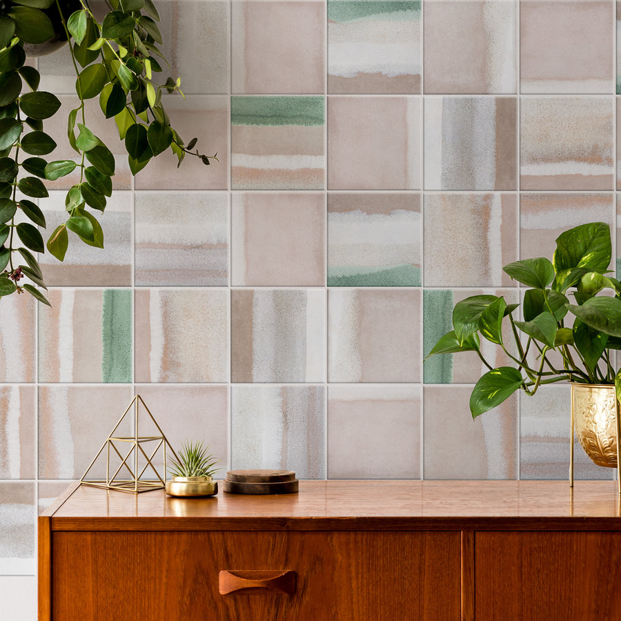 A tiled wall with a pattern of muted pink, beige, and green squares. A wooden sideboard holds green potted plants, a gold geometric decor piece, and small trays. Green hanging plants drape from above.