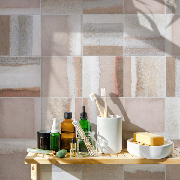 A bathroom shelf holds skincare bottles, jars, a jade roller, glass dropper bottles, toothbrushes in a holder, a bowl with soap bars, and a hairbrush, set against patterned tile walls in natural sunlight.