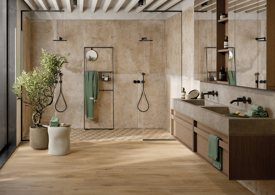 Modern bathroom with beige stone walls and wood accents, featuring two walk-in showers with black fixtures, double sinks, green towels, shelves with toiletries, a potted plant, and large windows letting in natural light.
