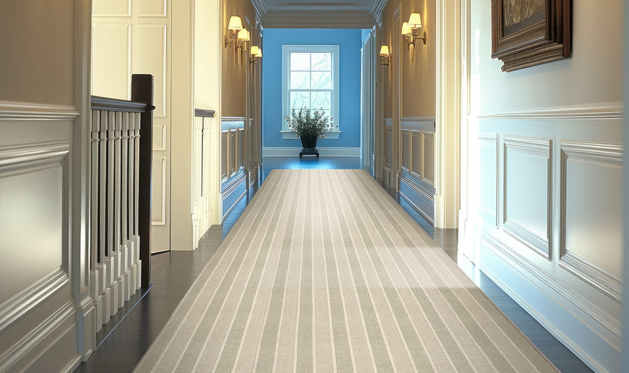 A long, elegant hallway with cream-colored walls, decorative molding, and soft lighting from wall sconces, featuring a striped beige runner rug and a potted plant placed near a window at the far end.