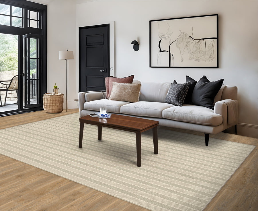 A modern living room with a light gray sofa, assorted pillows, a dark wood coffee table, beige striped rug, and wall art. The space has wooden floors, a black door, and glass doors leading outside.
