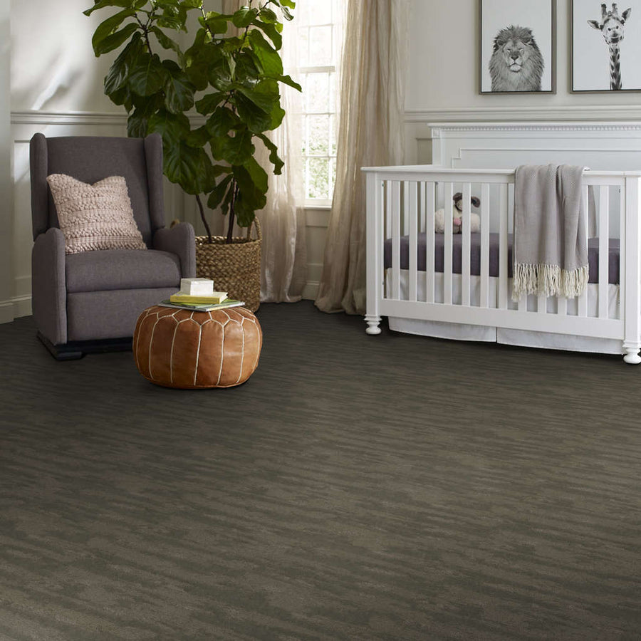A nursery with a gray crib, a gray armchair with a pink pillow, a brown leather ottoman, a large potted plant, and framed animal prints on the wall. Light flows in from a window with sheer curtains.