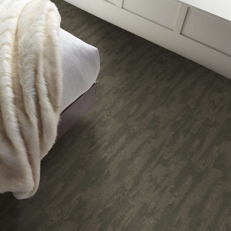 A bed with a white textured blanket and a cream-colored faux fur throw sits beside a dark, patterned carpet floor and a white paneled wall.