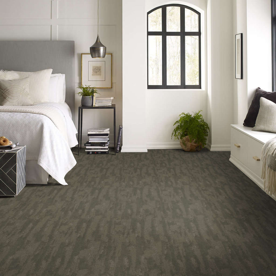Modern bedroom with gray carpet, a neatly made bed with white bedding, a black nightstand with decor, a large window with black trim, a potted plant, and a built-in bench with cushions and pillows.