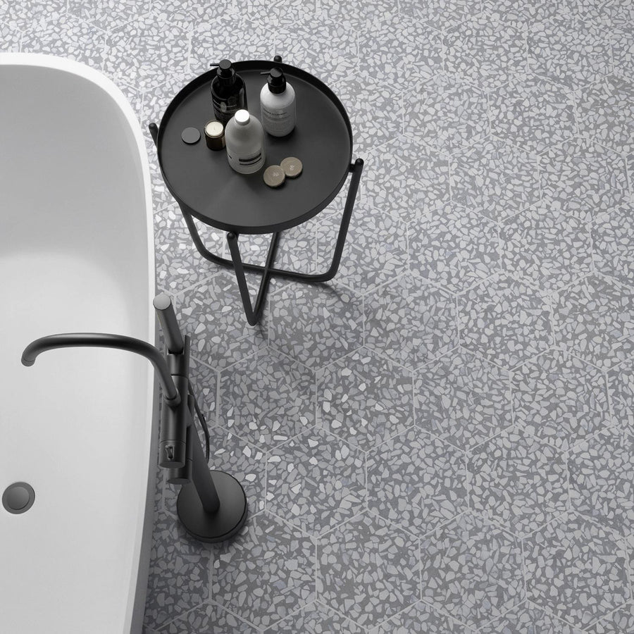 A modern bathroom with a white bathtub, a sleek silver faucet, and a black round side table holding toiletries. The floor is covered in gray terrazzo tiles with white and light gray patterns.