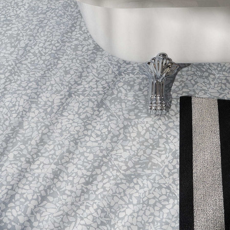 A close-up of a bathroom floor with light gray and white terrazzo tiles, a white bathtub, a silver faucet, and part of a black and white striped bath mat.