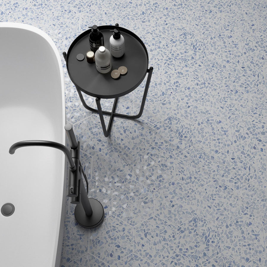 A modern bathroom with a white bathtub, a sleek silver faucet, and a small round black table holding toiletries, set on a light blue and white terrazzo floor.