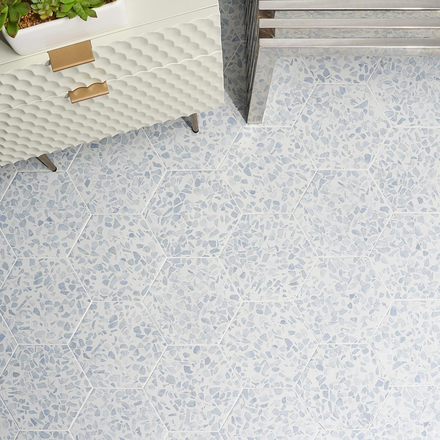 Hexagonal tiles with a terrazzo pattern in light blue and white cover the floor. Part of a textured white cabinet with gold handles and a metal bench are visible in the corner.