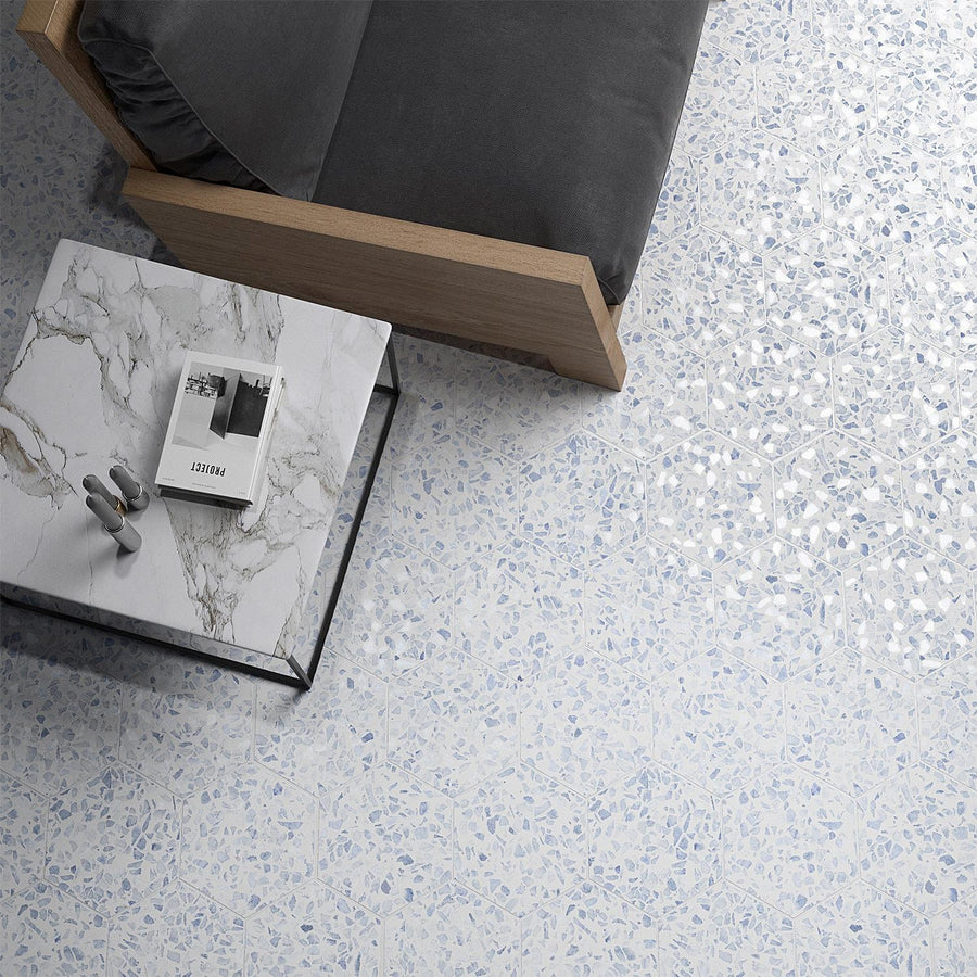 A top-down view of a light terrazzo floor, a gray cushioned wooden chair, and a marble coffee table with a magazine and a decor item on it.