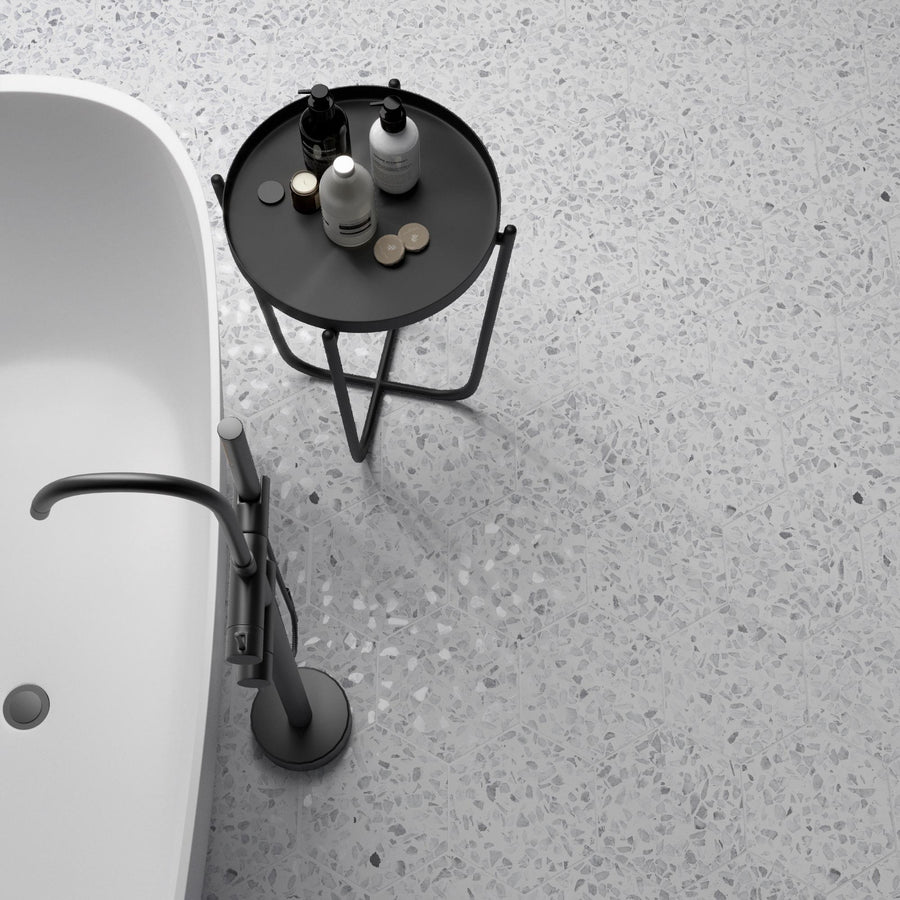 A modern bathroom with a freestanding white bathtub, a sleek silver faucet, and a round black side table holding various toiletries on a light terrazzo floor.